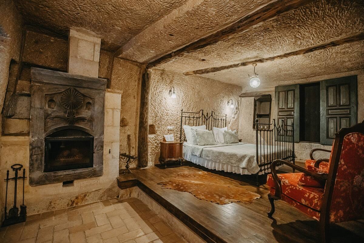 Una habitación rústica con chimenea de piedra, cama con ropa de cama blanca, un banco de madera y un tapiz de piel de vaca.