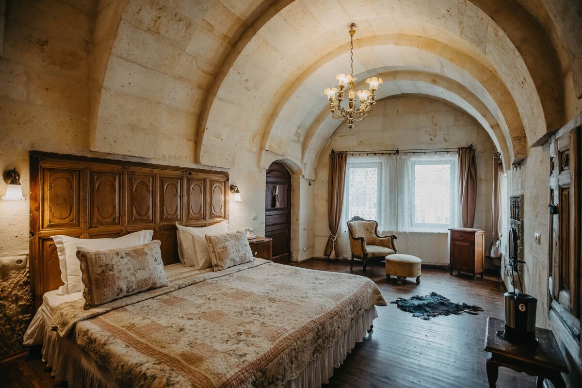 Un dormitorio con una cama grande, cabecero de madera, techo abovedado, candelabro y vistas al exterior a través de una ventana.