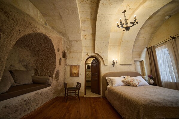 A room with a stone wall, a wooden bench, a bed with pillows, a wooden chair, and a chandelier. - Meleklerevi Cave Hotel (Ürgüp)