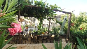 A stone archway leading to a covered outdoor seating area with metal chairs and tables, surrounded by lush greenery and flowering plants.