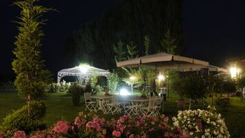 Un jardín al aire libre con un pabellón metálico blanco, una mesa y sillas cubiertas, y diversas plantas y flores en macetas.