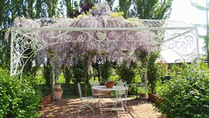 A garden with a white metal pergola covered in purple flowers, a round table with chairs, and potted plants. - Agriturismo Millefiori Corte delle Rose (Rovigo)