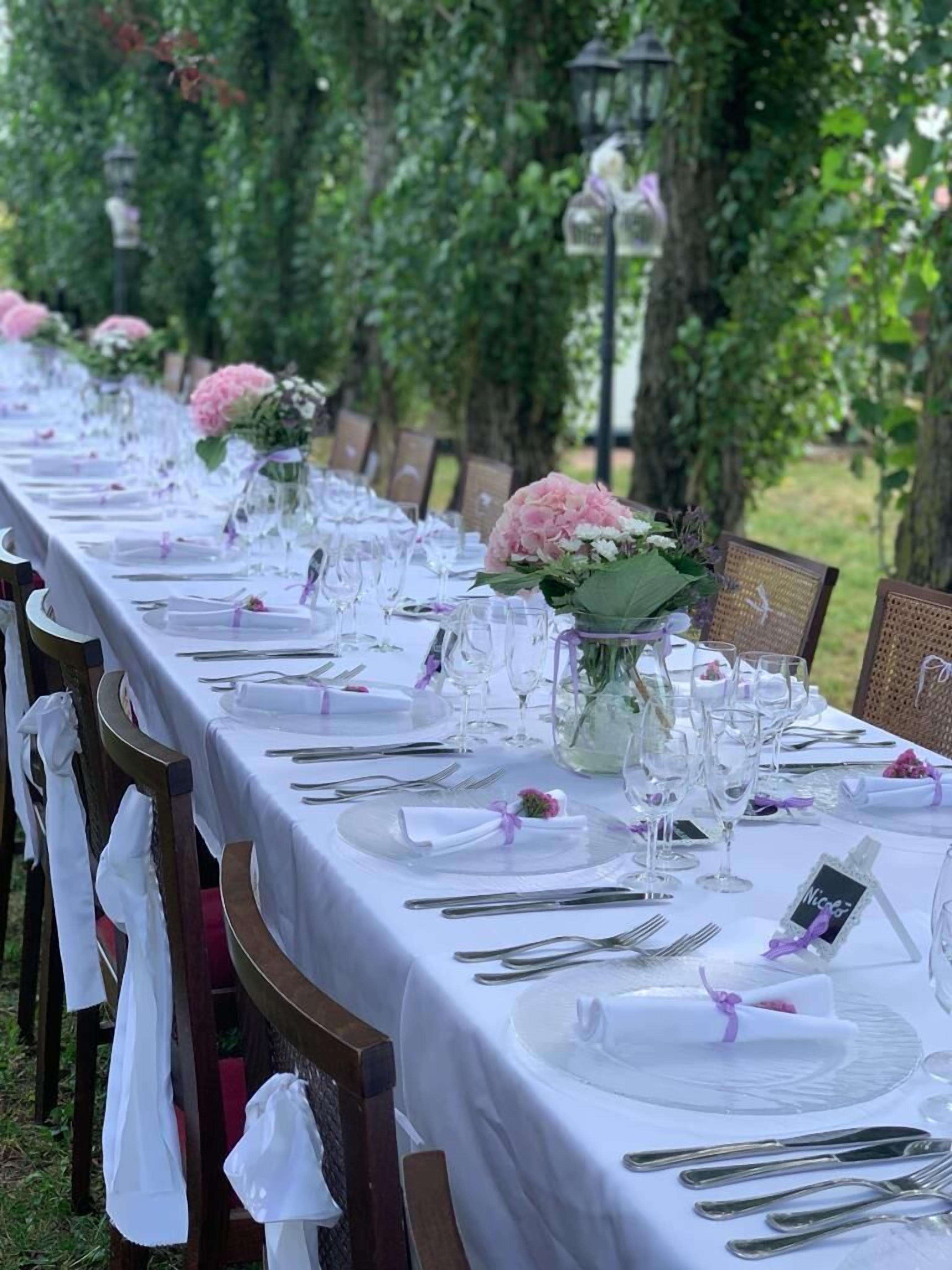 Ein langer Esstisch im Freien, gedeckt mit weißen Tischdecken, Gläsern und floralen Tischdekorationen.