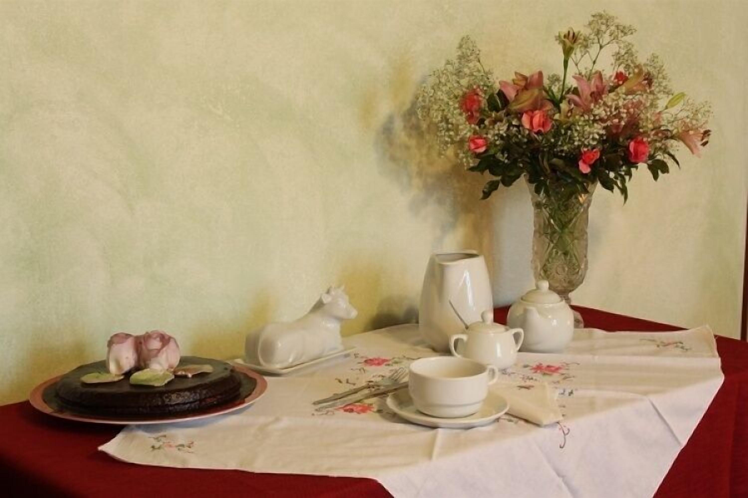 Una mesa dispuesta con un pastel, tetera, tazas, platillos y un jarrón de flores.