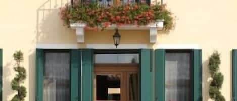 Un edificio con balcón decorado con flores, una puerta de madera y contraventanas verdes.