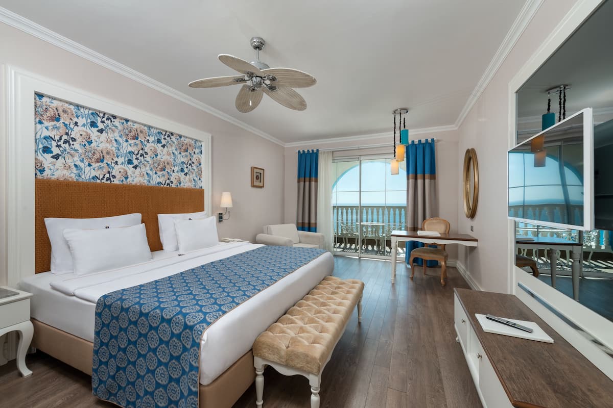 A hotel room with a large bed, a desk, a chair, a bench, a ceiling fan, and a large window with curtains.
