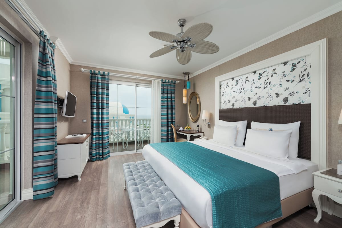 A hotel room with a large bed, a ceiling fan, a balcony with curtains, a desk, and a bench.