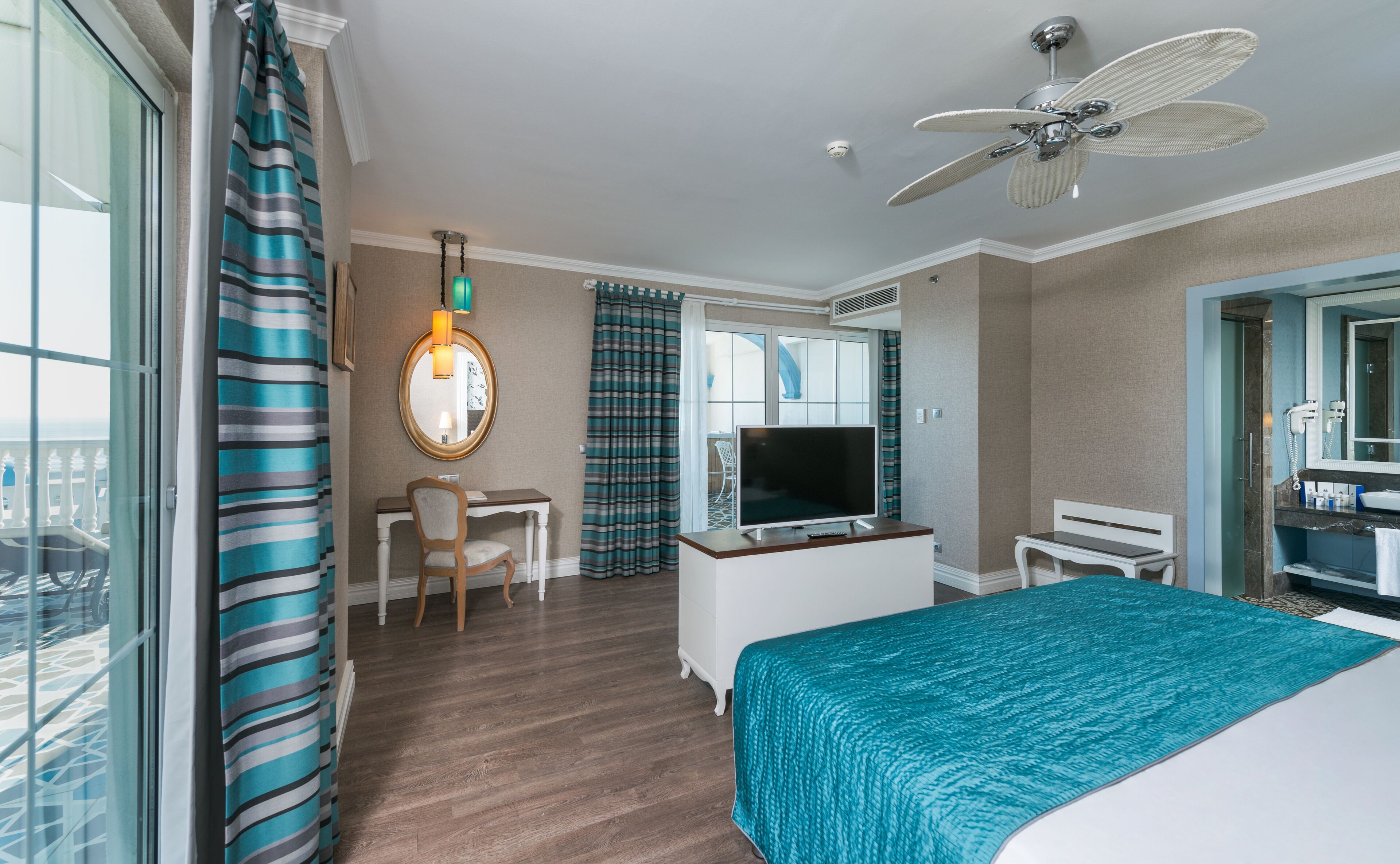 A hotel room with a bed, a desk, a chair, a TV, a ceiling fan, and a bathroom with a mirror and a hairdryer.