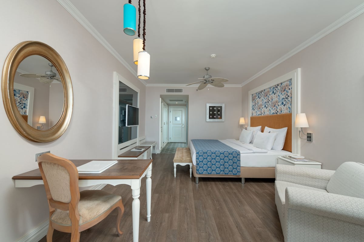 A hotel room with a wooden desk, a chair, a bed with a patterned bedspread, a nightstand with a lamp, a wall-mounted mirror, and a ceiling fan.