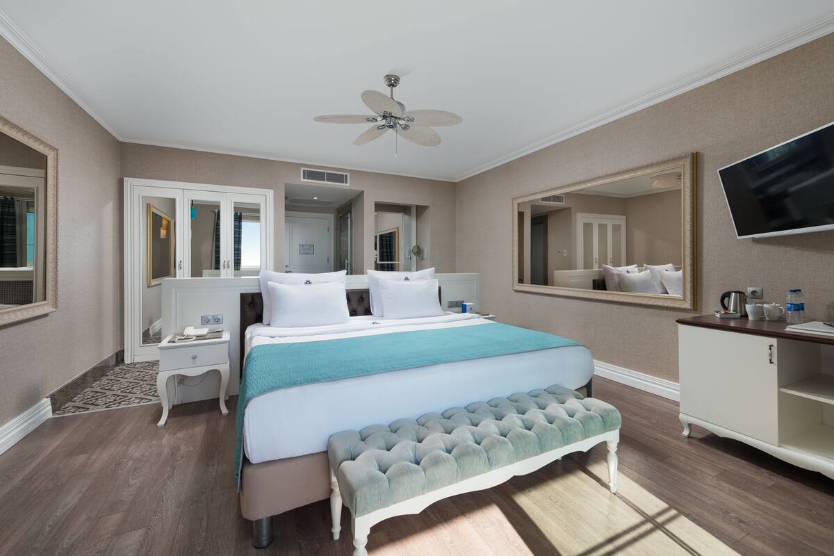 A hotel room with a large bed, a bench, a desk, a TV, and a mirror.