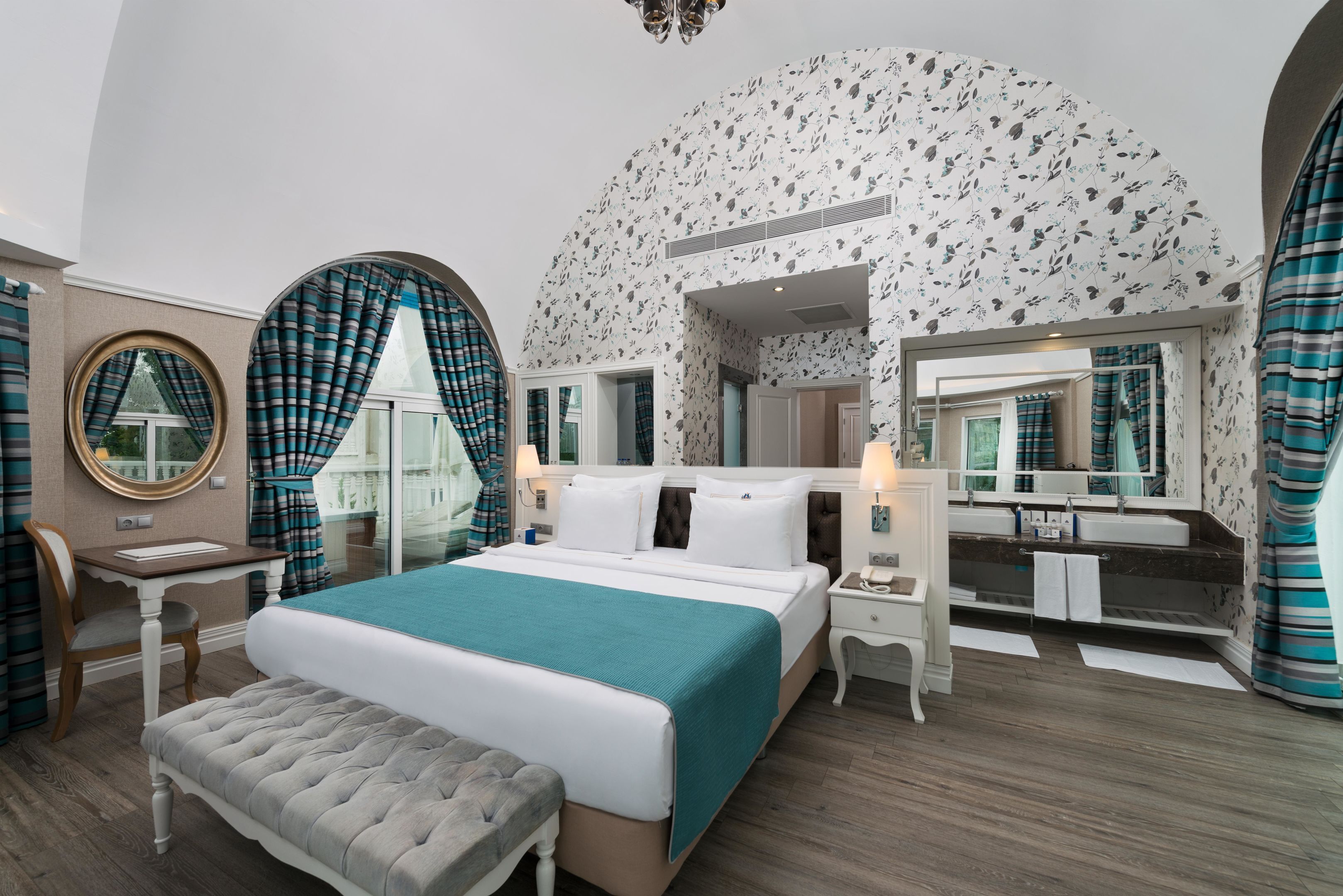 A bedroom with a large bed, a bedside table, a chair, a bench, a mirror, and a bathroom with a sink and mirror.