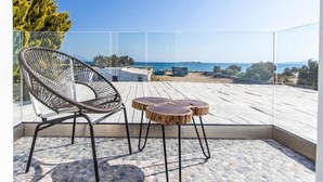 A balcony with a wicker chair and two round wooden tables, overlooking a beach and ocean. - Evdokia (Naxos)
