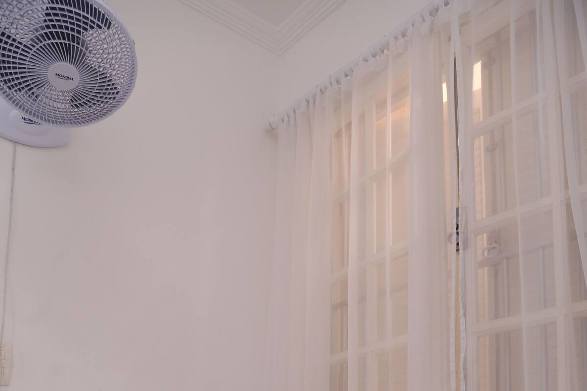 A white room with a bed, a window with sheer curtains, and a standing fan.