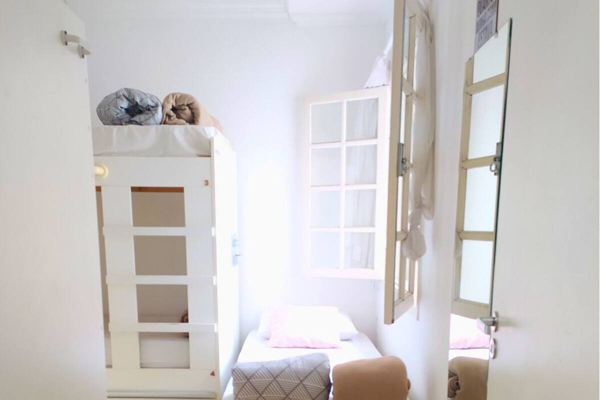 A compact bedroom with a bed, a small stool, a window, and a door.