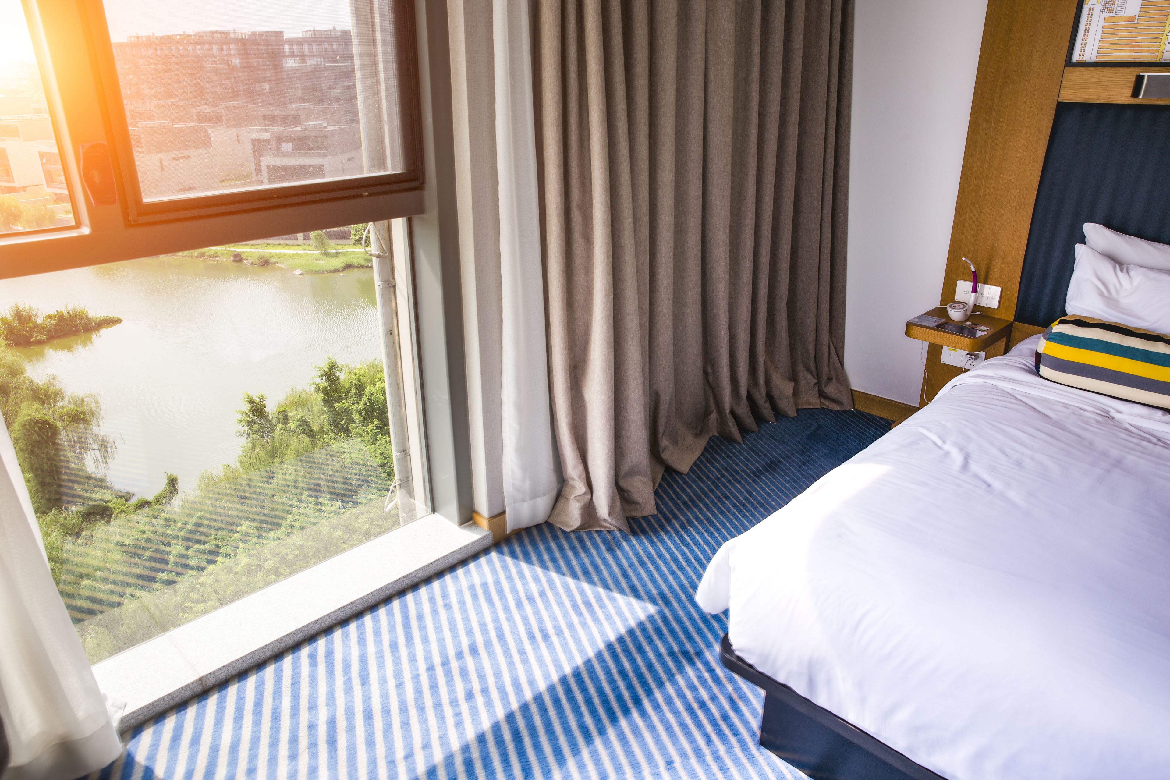 A hotel room with a large window, a bed with a striped pillow, a nightstand with a lamp, and a view of a body of water and greenery.