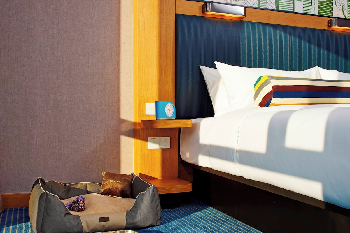 Een hotelkamer met een bed, nachtkastje met klok, klein schap en een ruimte voor bagage.