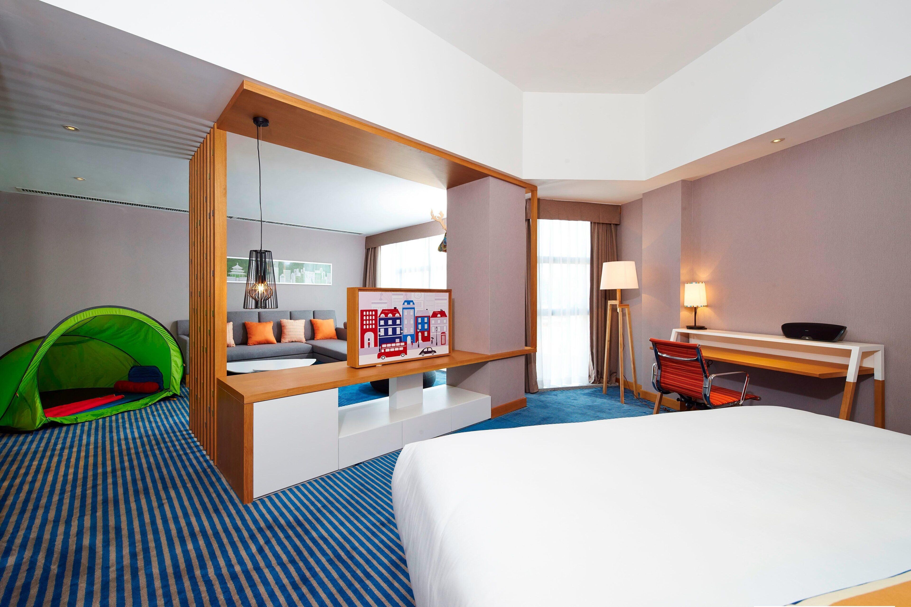 Habitación de hotel con cama, escritorio, silla, lámpara, una tienda de campaña verde y un cuadro en la pared.