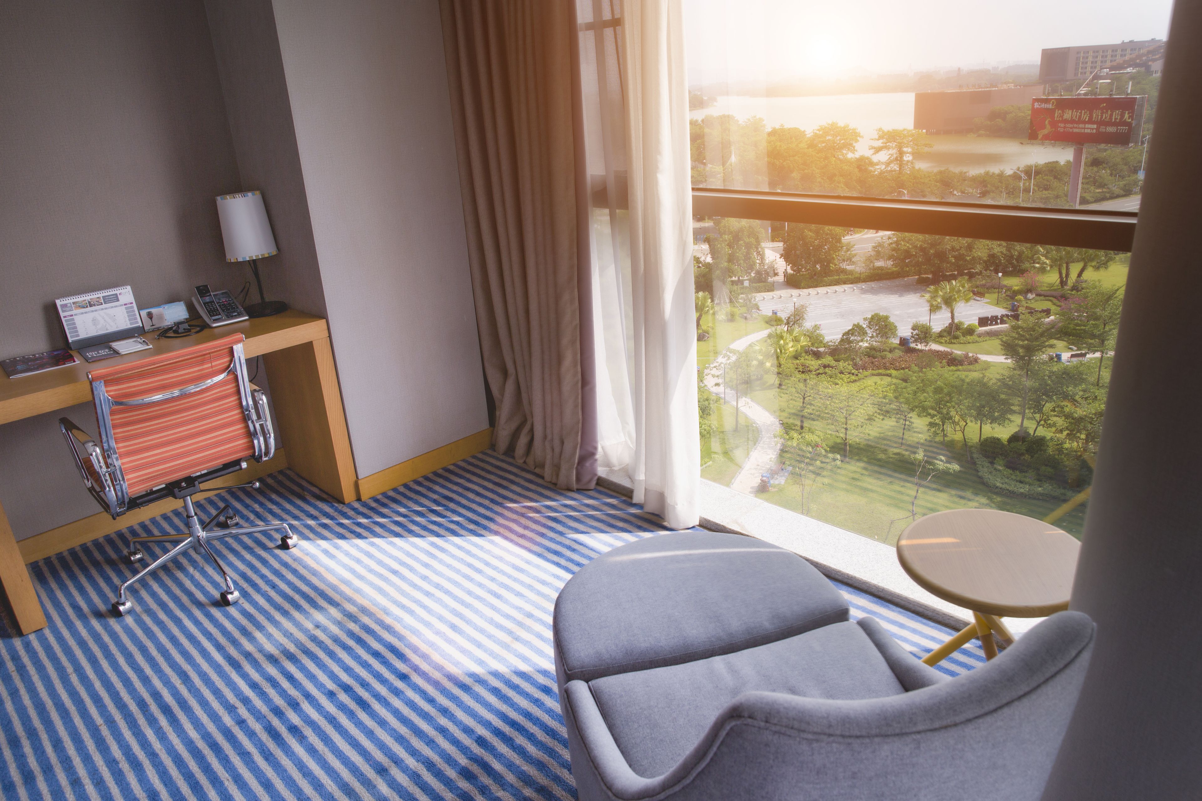 A hotel room with a desk, chair, and a view of a park and buildings.