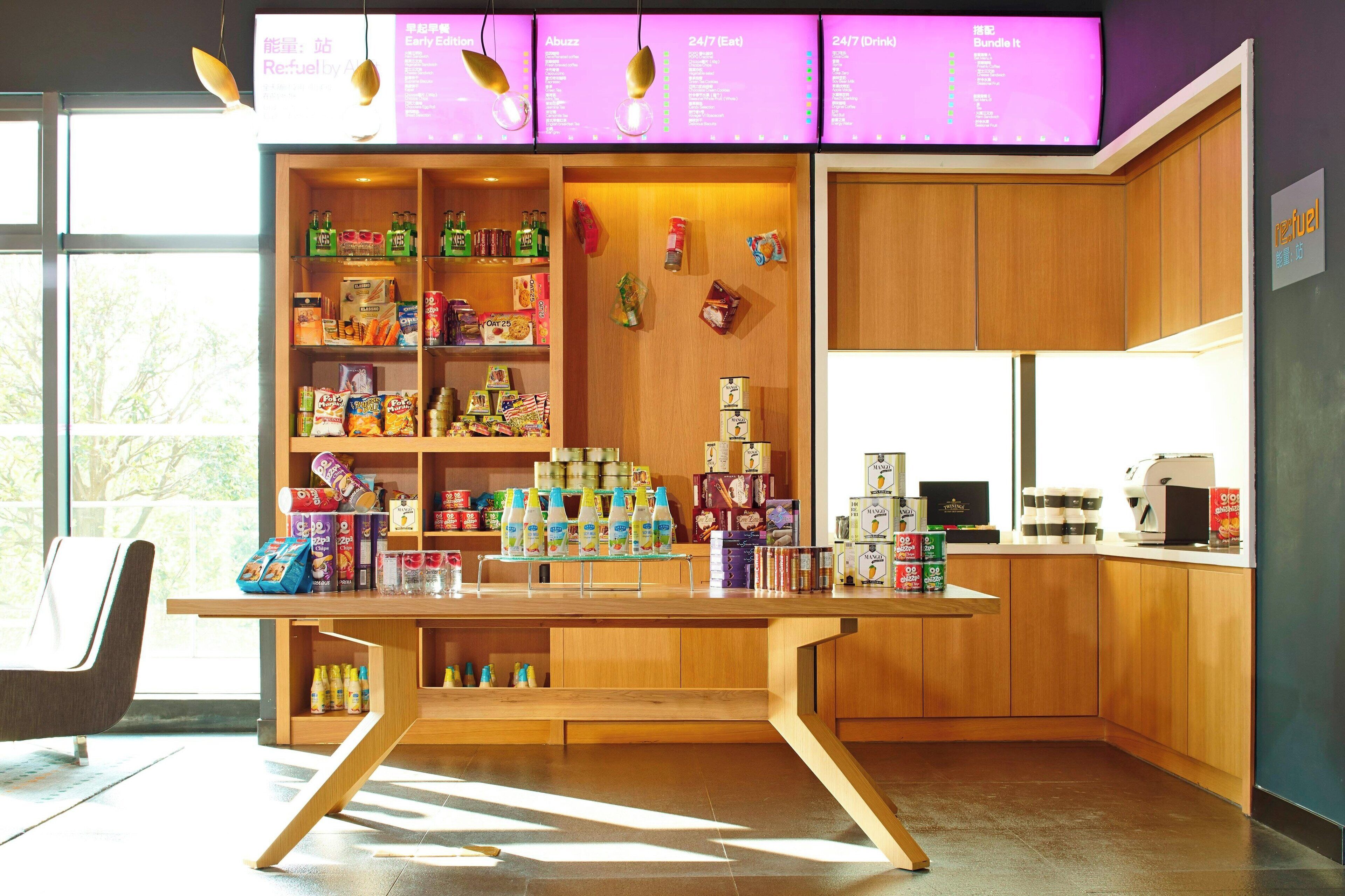 Una cafetería moderna con un mostrador de madera, estantes llenos de aperitivos y bebidas, y un panel con la carta.
