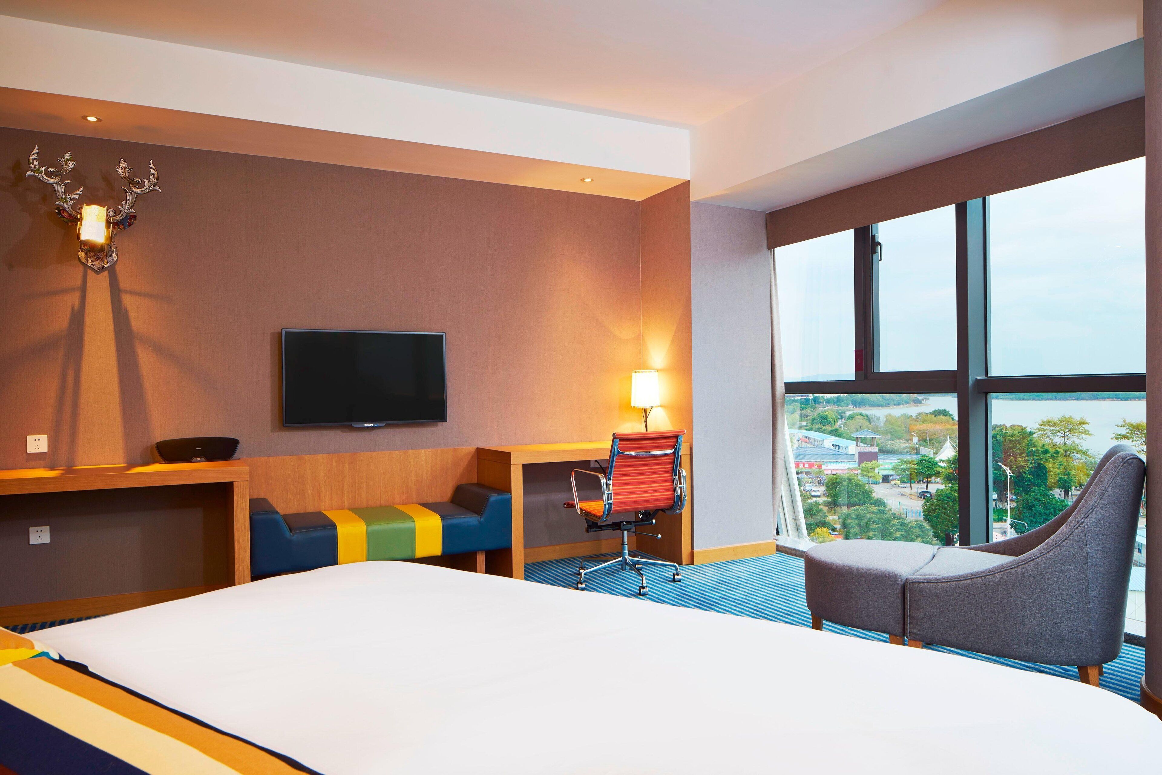 A hotel room with a bed, a television, a desk with a lamp, a chair, and a view of the city through large windows.
