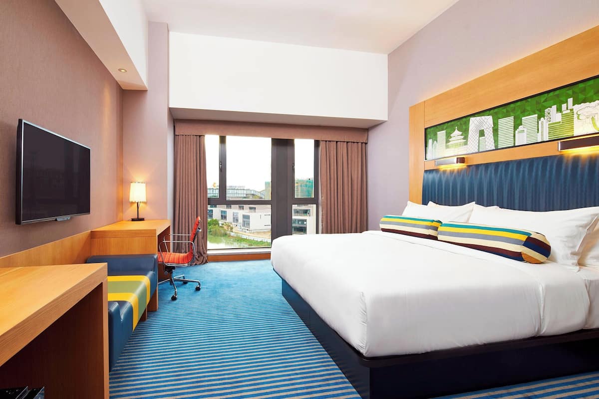 Een hotelkamer met een groot bed, een bureau met stoel, een televisie en uitzicht op de stad door grote ramen.