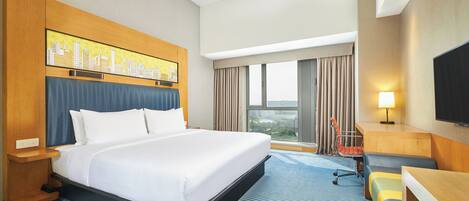 A hotel room with a large bed, a desk, a chair, a TV, and a cityscape artwork on the wall.