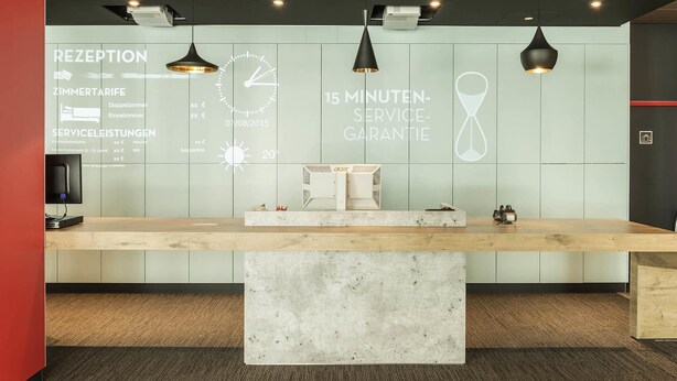 A reception desk with a digital display, pendant lighting, and a marble counter. - Ibis Muenchen City Ost (Munich)