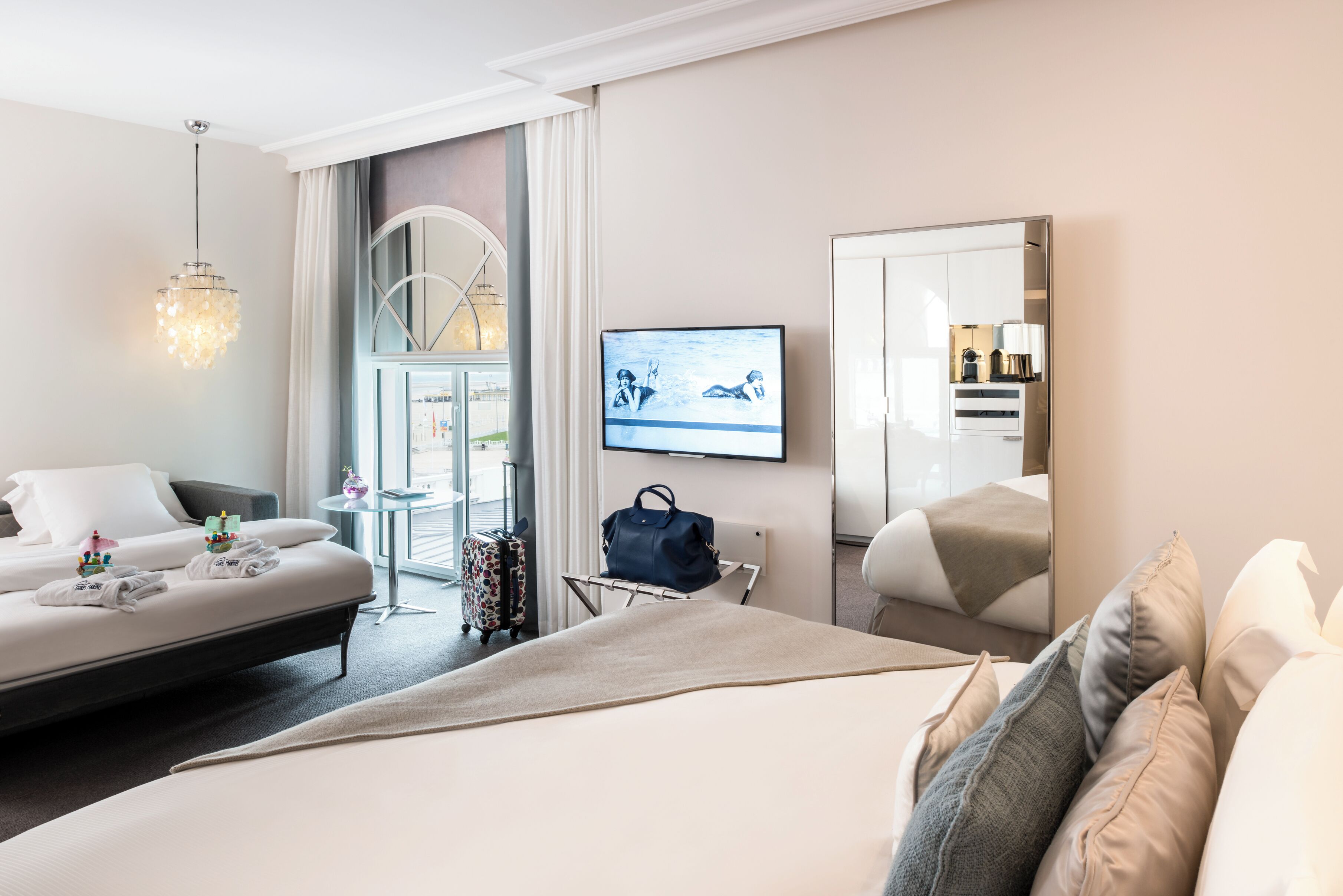 A hotel room with a bed, a seating area with a sofa, a television, a suitcase, and a mirror.