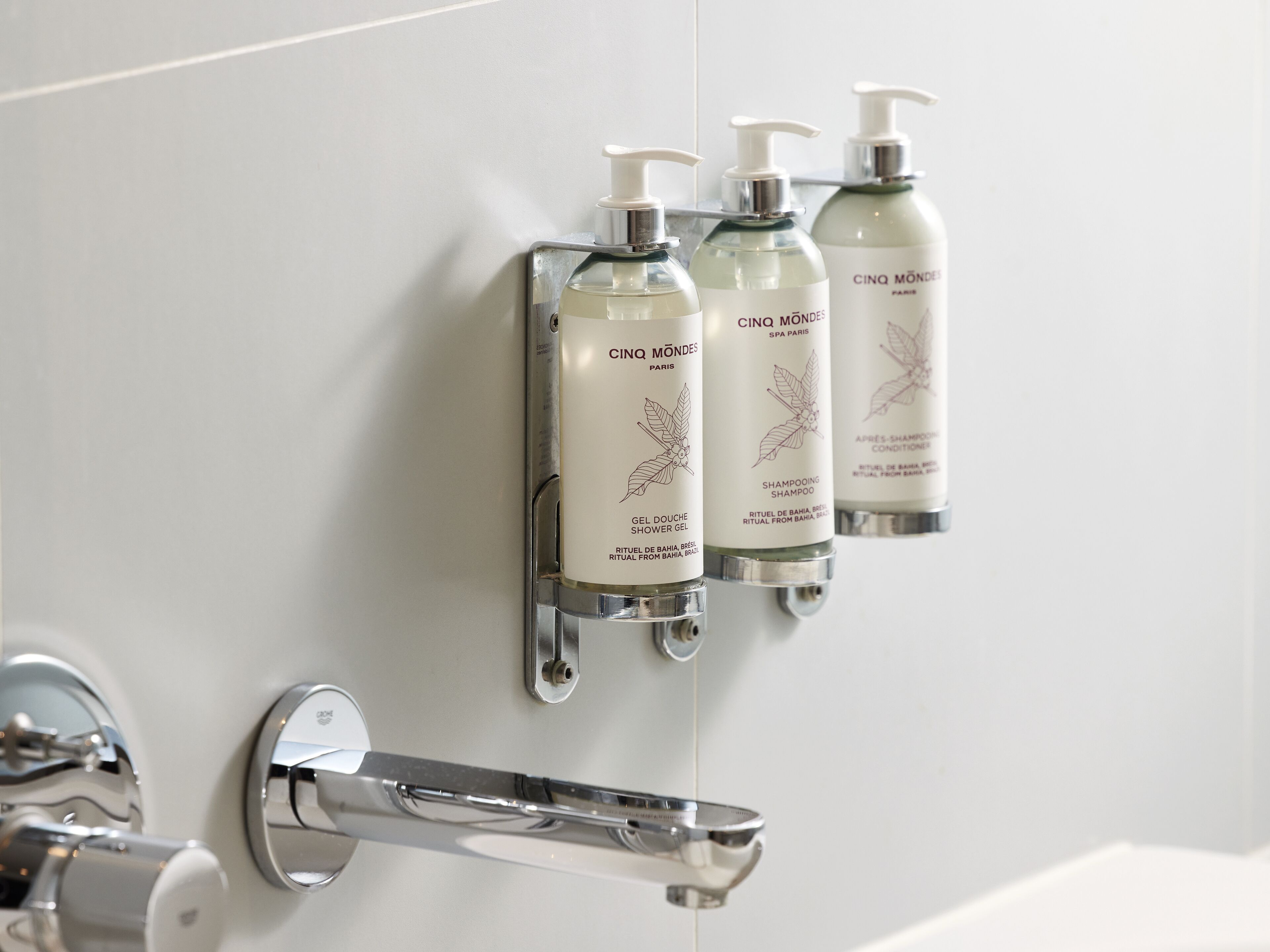 A bathroom with three bottles of Cinq Mondes shampoo and shower gel mounted on a wall.