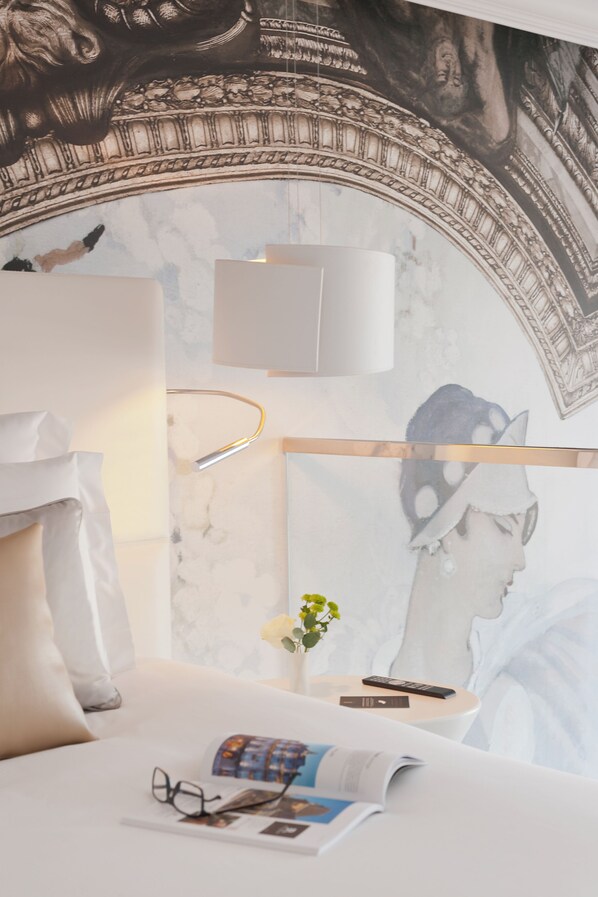 Un dormitorio con pared de mármol, una cama con ropa de cama blanca, una mesita de noche con lámpara, un control remoto y una revista.