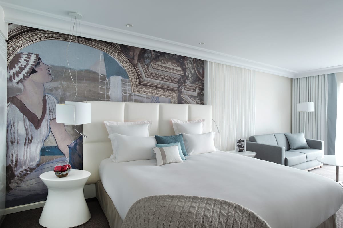 Une chambre d’hôtel moderne avec un grand lit, un canapé gris et une fresque murale décorative.