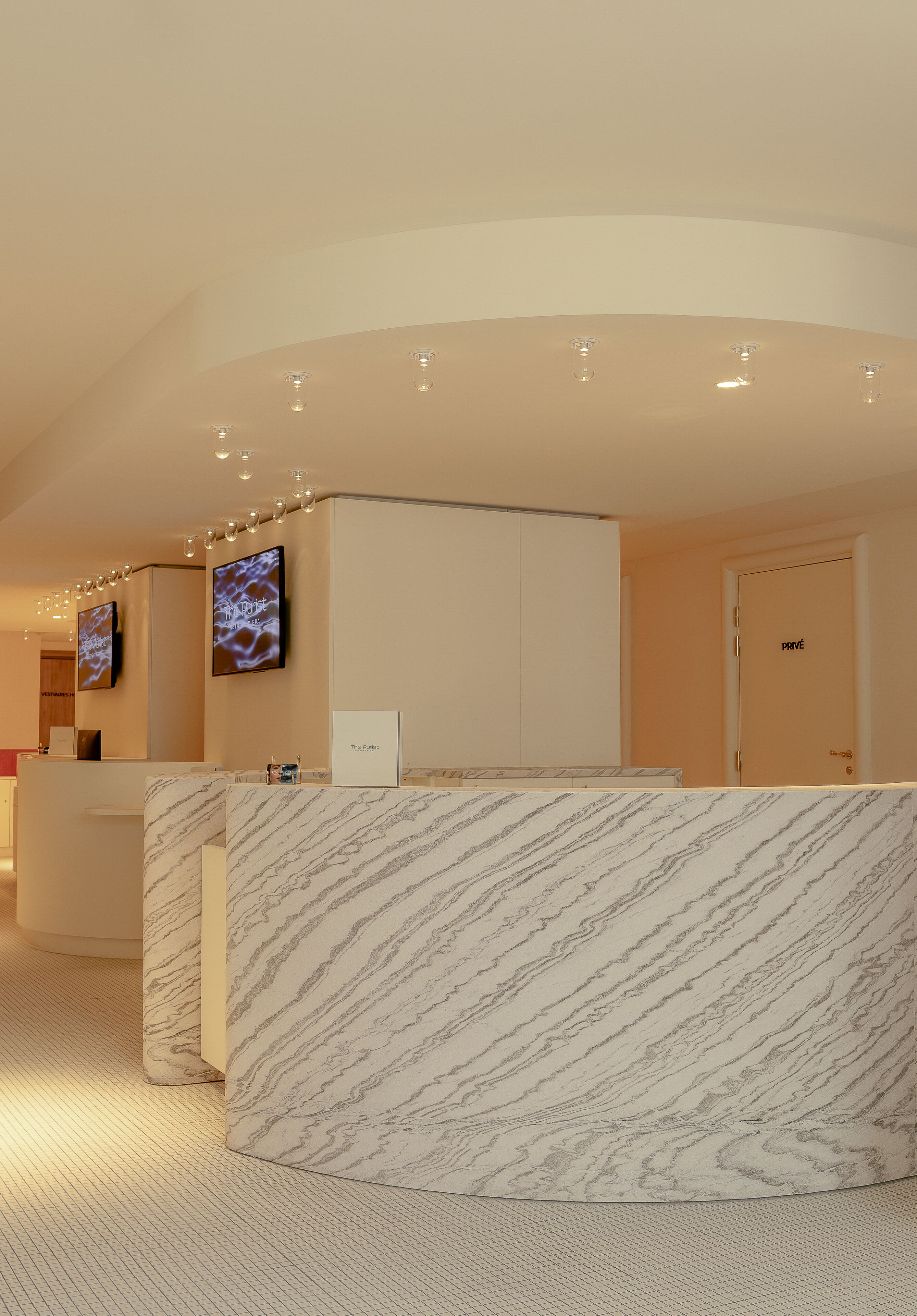 Een moderne hotellobby met marmeren receptiebalies, een receptie en een televisiescherm, met een deur waarop 'POVÉE' staat.