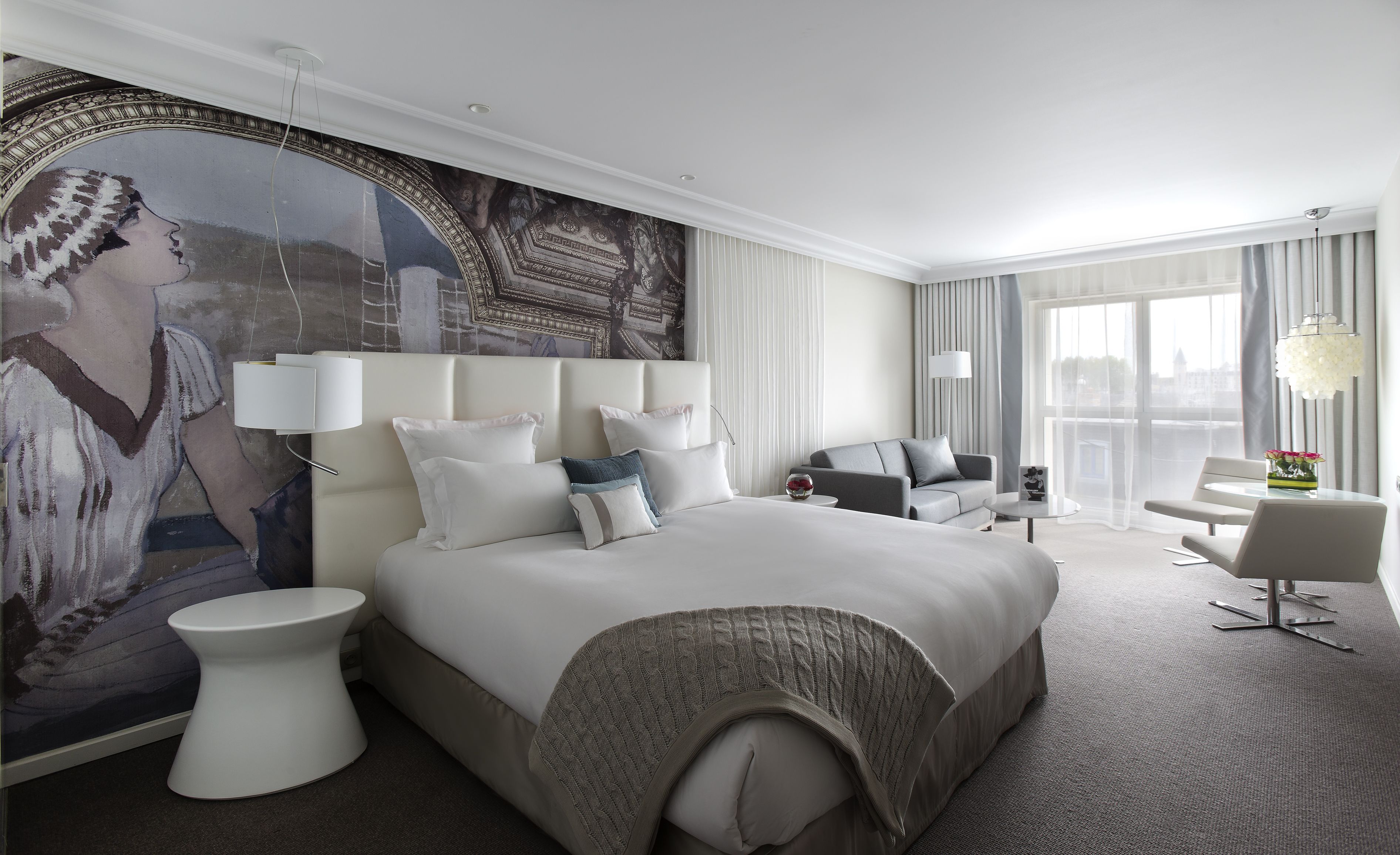 A modern hotel room with a large bed, a seating area with a sofa and armchairs, a small table, and a large window with curtains.