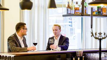 Deux personnes sont assises au bar, chacune avec un verre de vin à la main. On aperçoit en arrière-plan une étagère à bouteilles et des lampes suspendues.