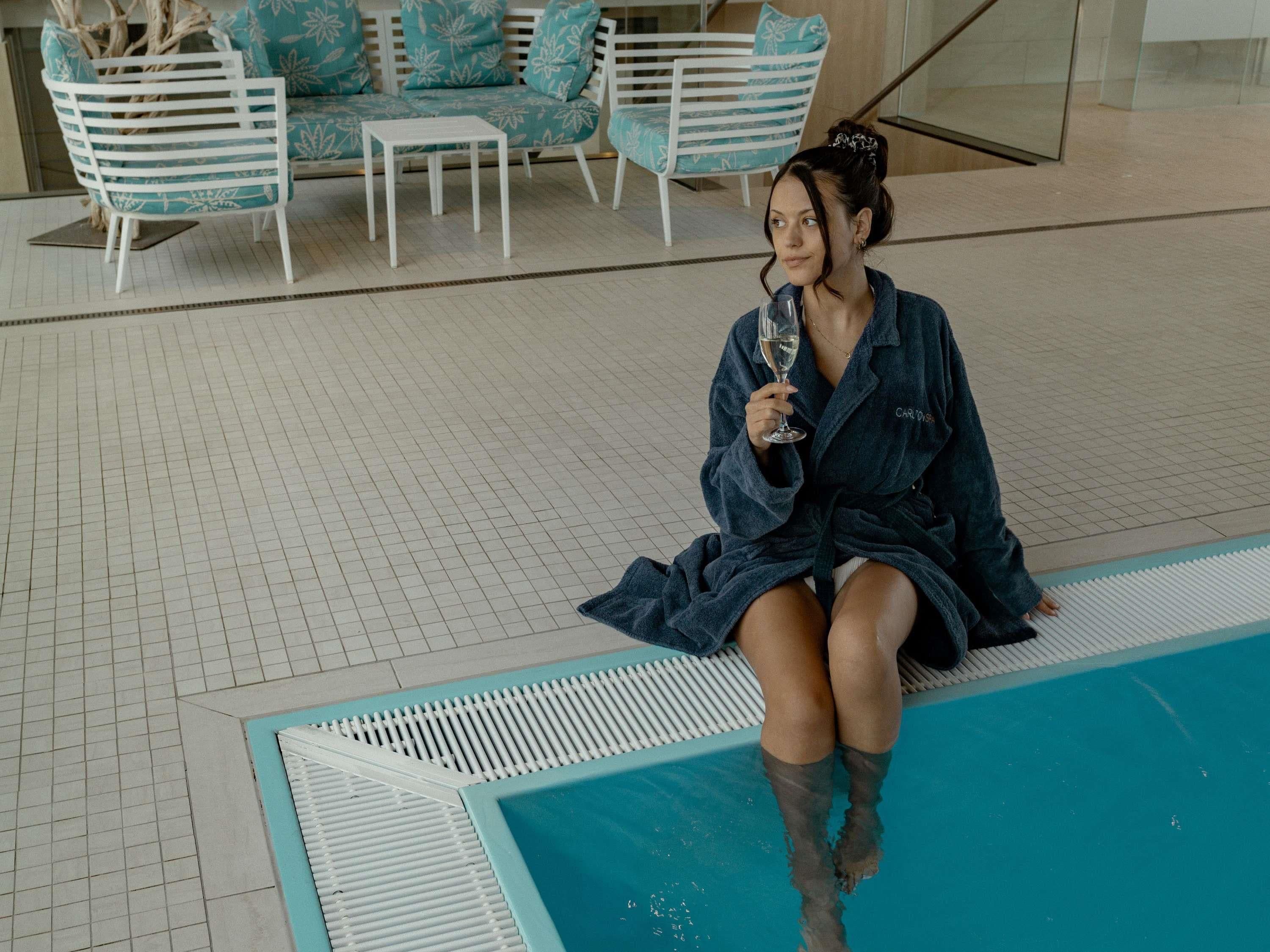 A person sitting by a pool with a glass of wine, surrounded by lounge chairs and a sofa with floral patterns.