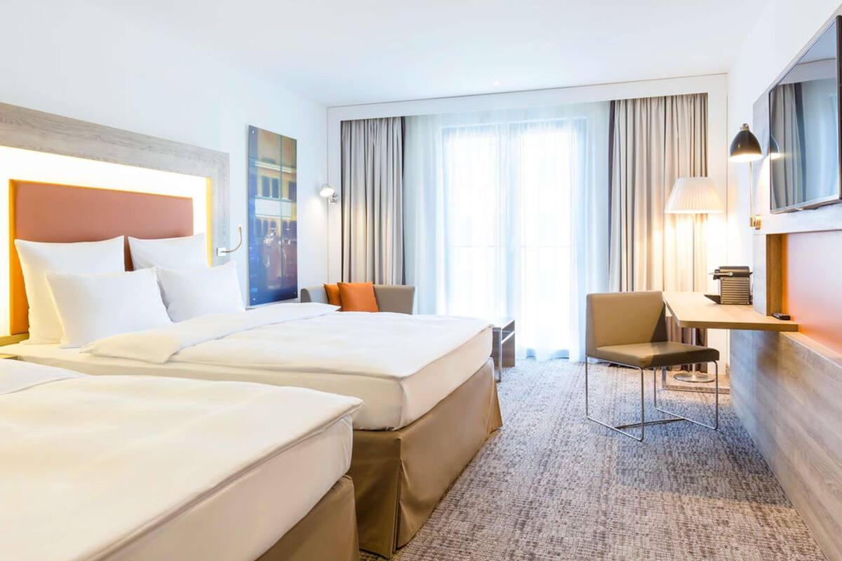 A hotel room with two beds, a desk, a chair, a TV, and a large window with curtains.