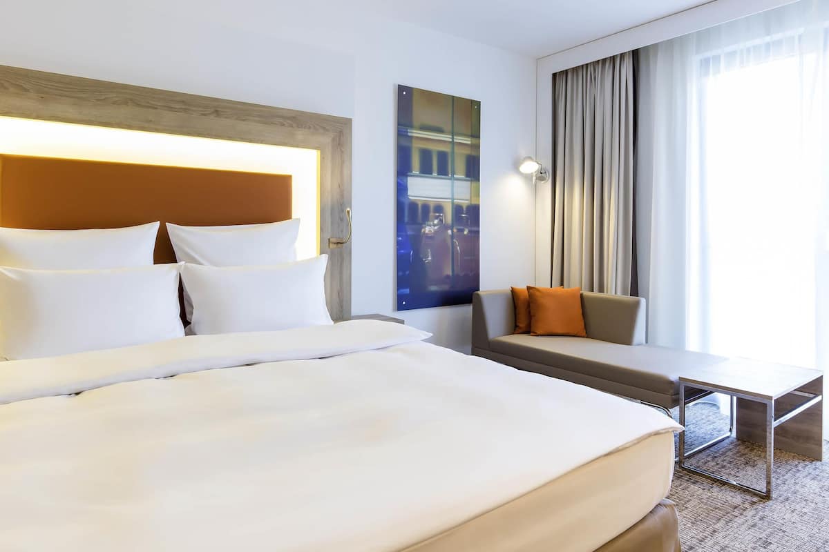 A hotel room with a large bed, a sofa, a small table, and a large window with curtains.