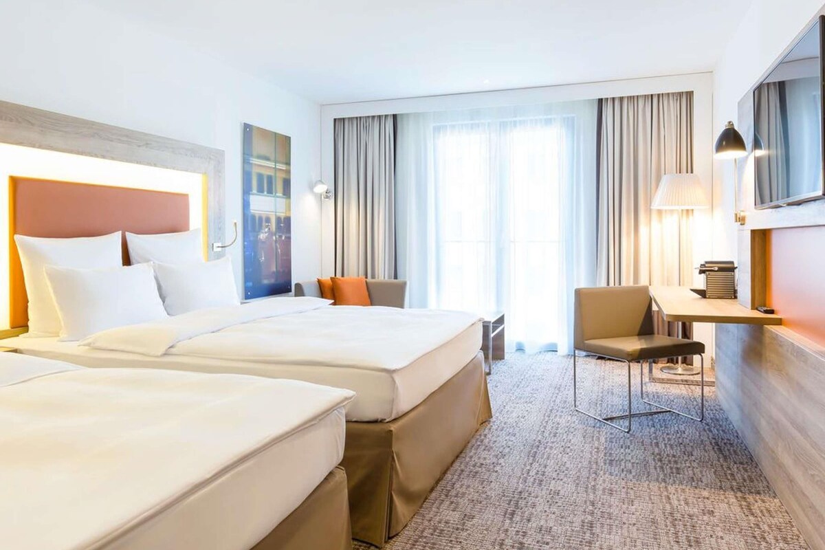 A hotel room with two beds, a desk, a chair, and a large window with curtains.