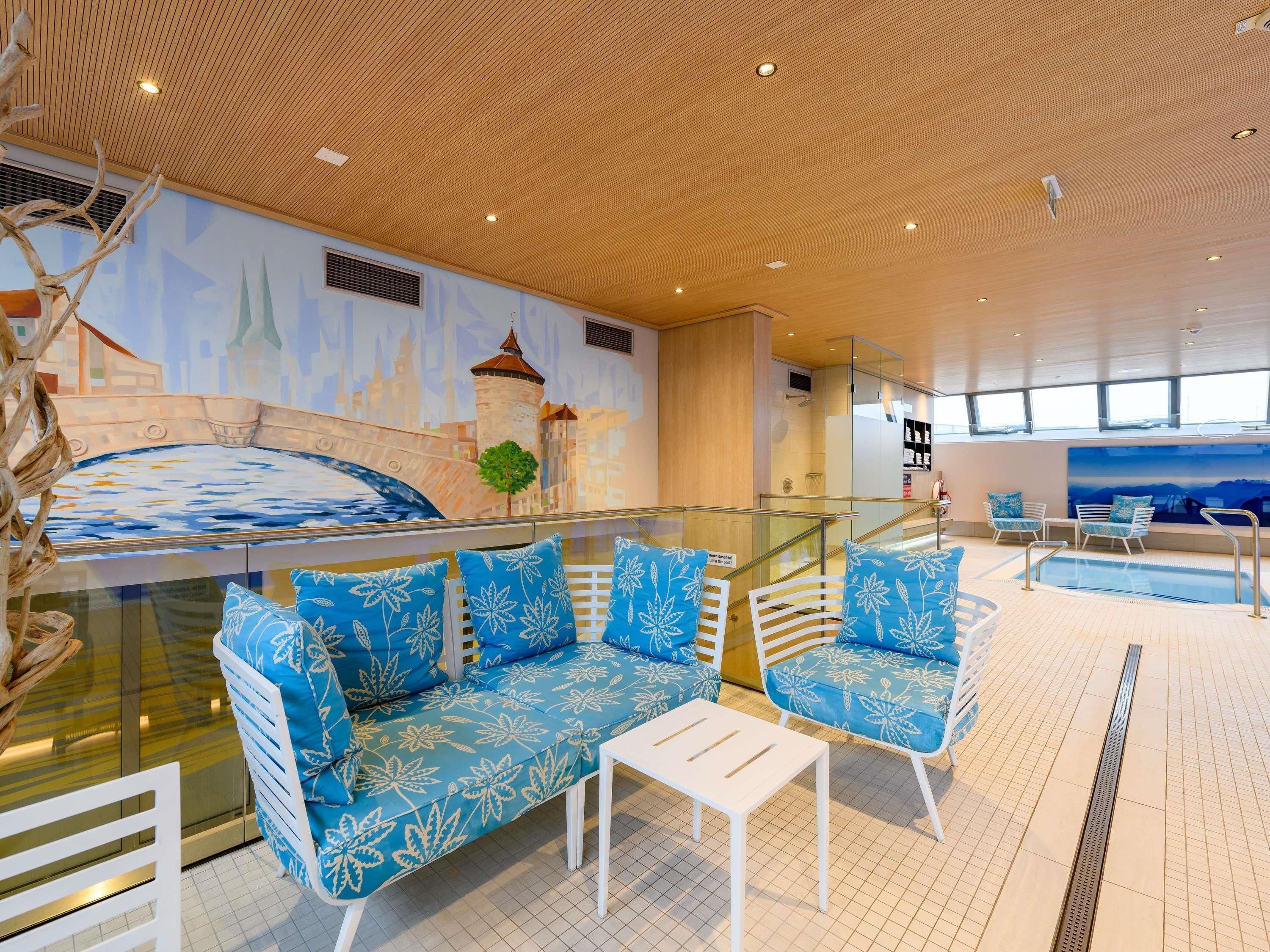 A modern indoor pool area with a sofa and chairs, a glass railing, and a mural of a bridge.