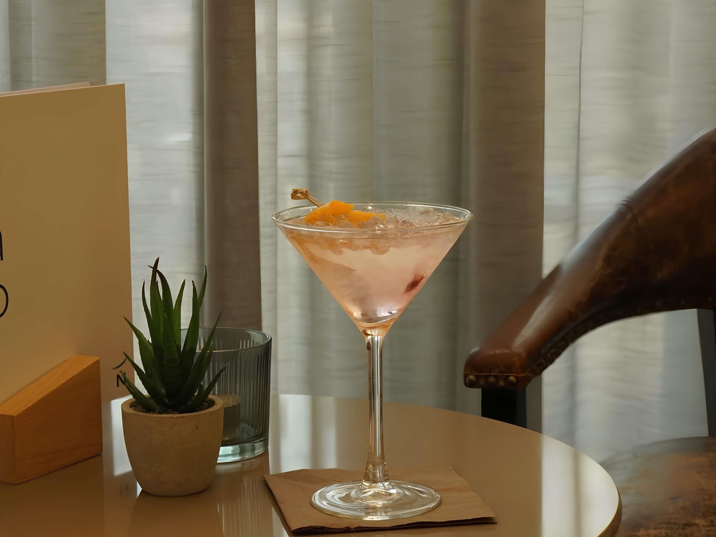A martini glass with an orange garnish on a round table, a small potted plant, and a glass of water.