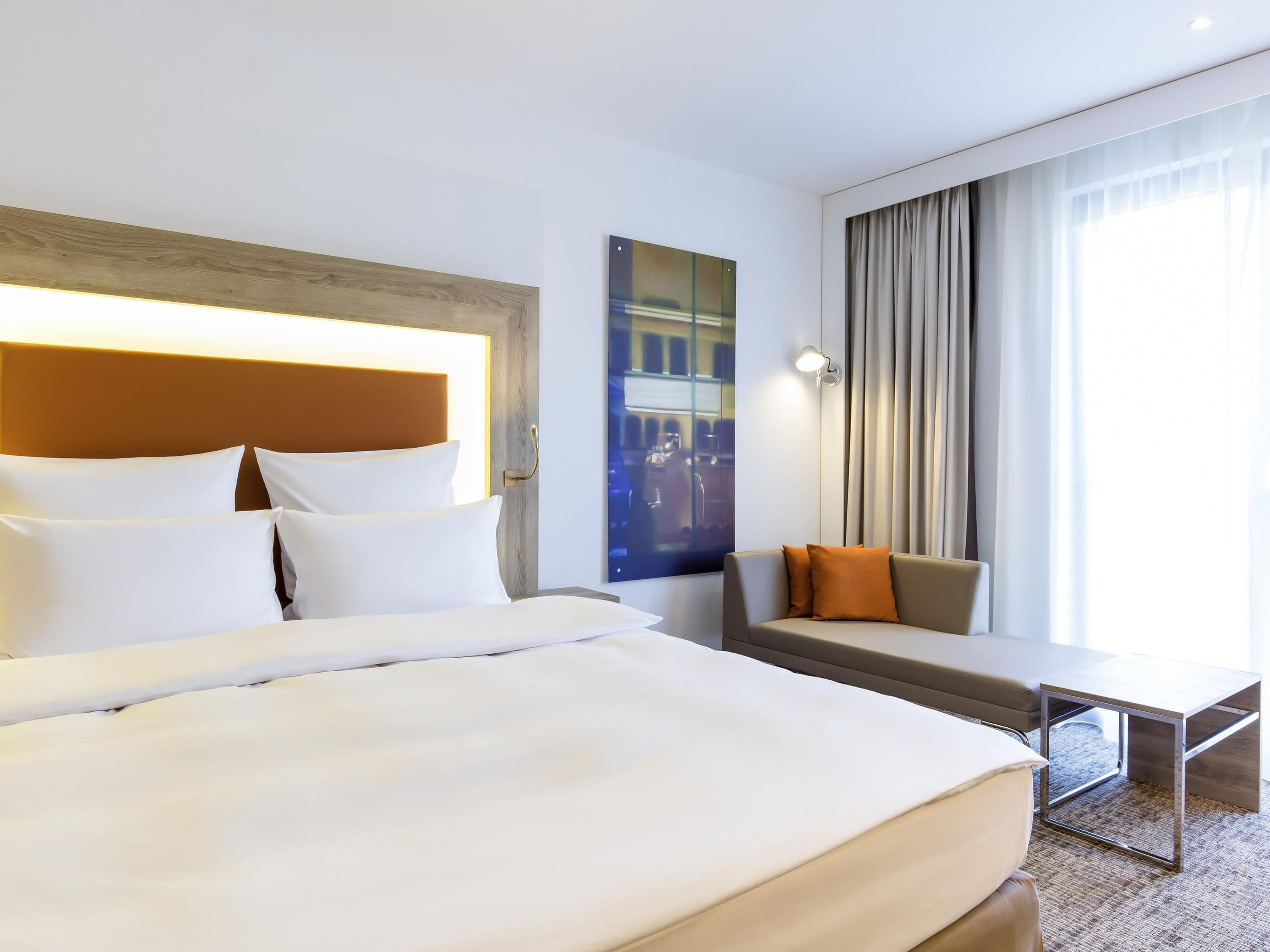 A hotel room with a bed, a sofa, a small table, and a large window with curtains.