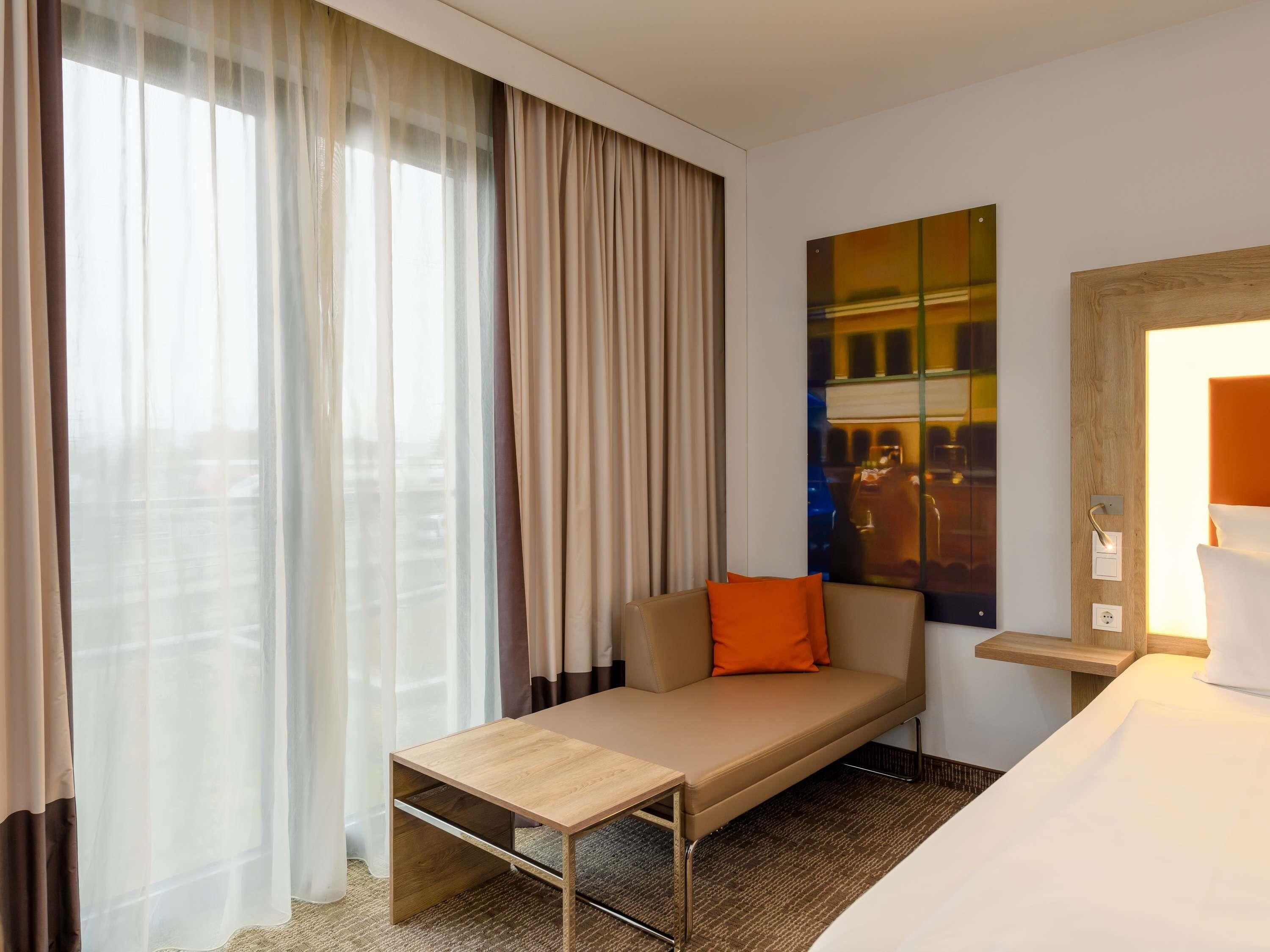 A hotel room with a bed, a sofa, a small table, a painting, and a window with curtains.