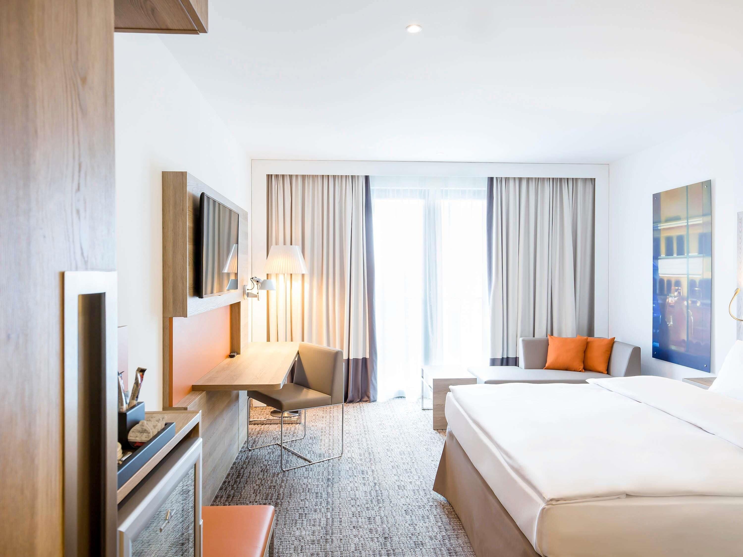 Habitación de hotel con una cama, un escritorio con silla, un televisor y una ventana con cortinas.