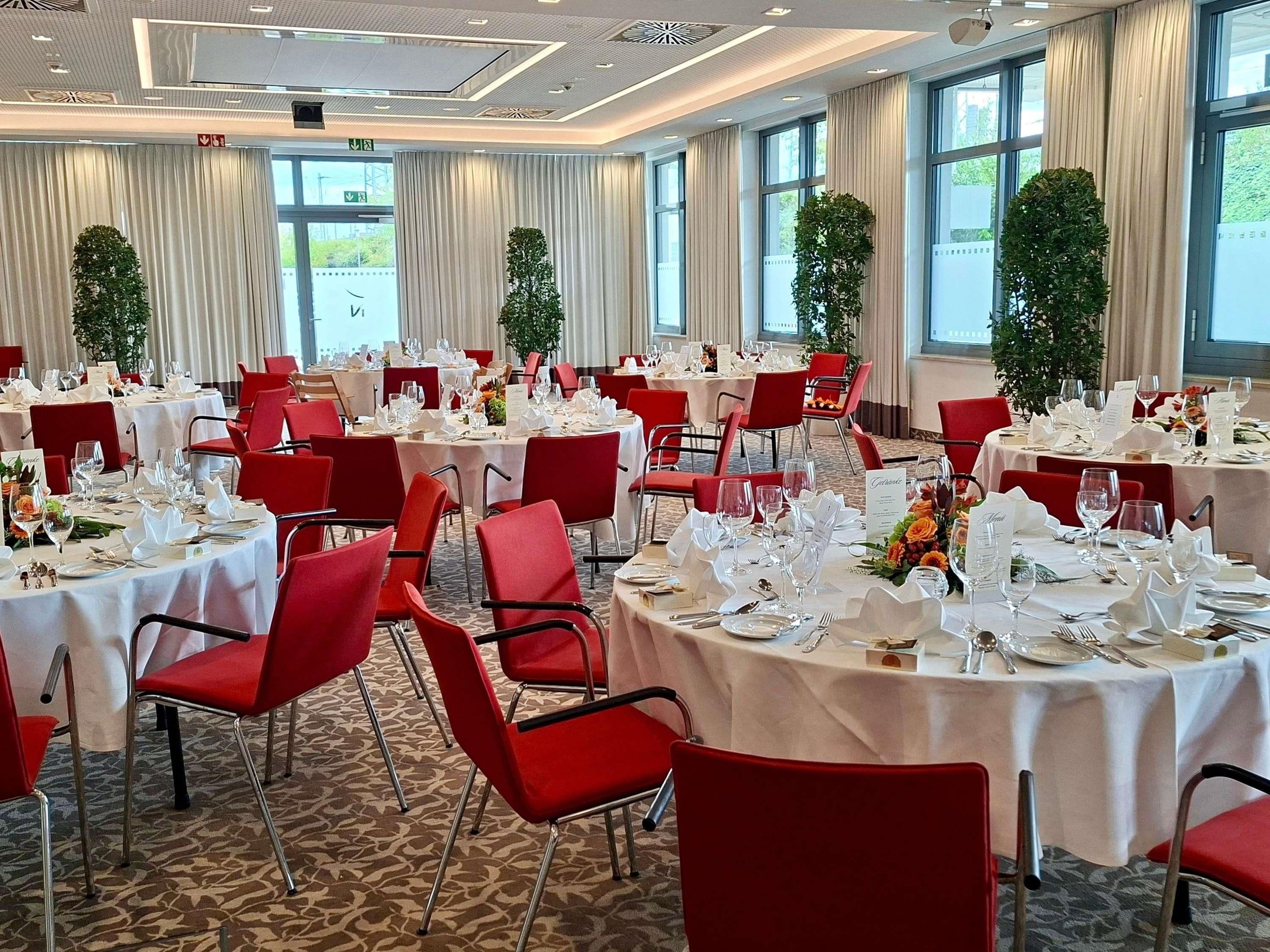 A banquet hall with round tables set for dining, red chairs, and large windows with curtains.