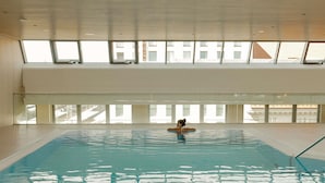 An indoor swimming pool with clear blue water, a person swimming, and large windows providing natural light. - Novotel Nuernberg Centre Ville (Nuremberg)