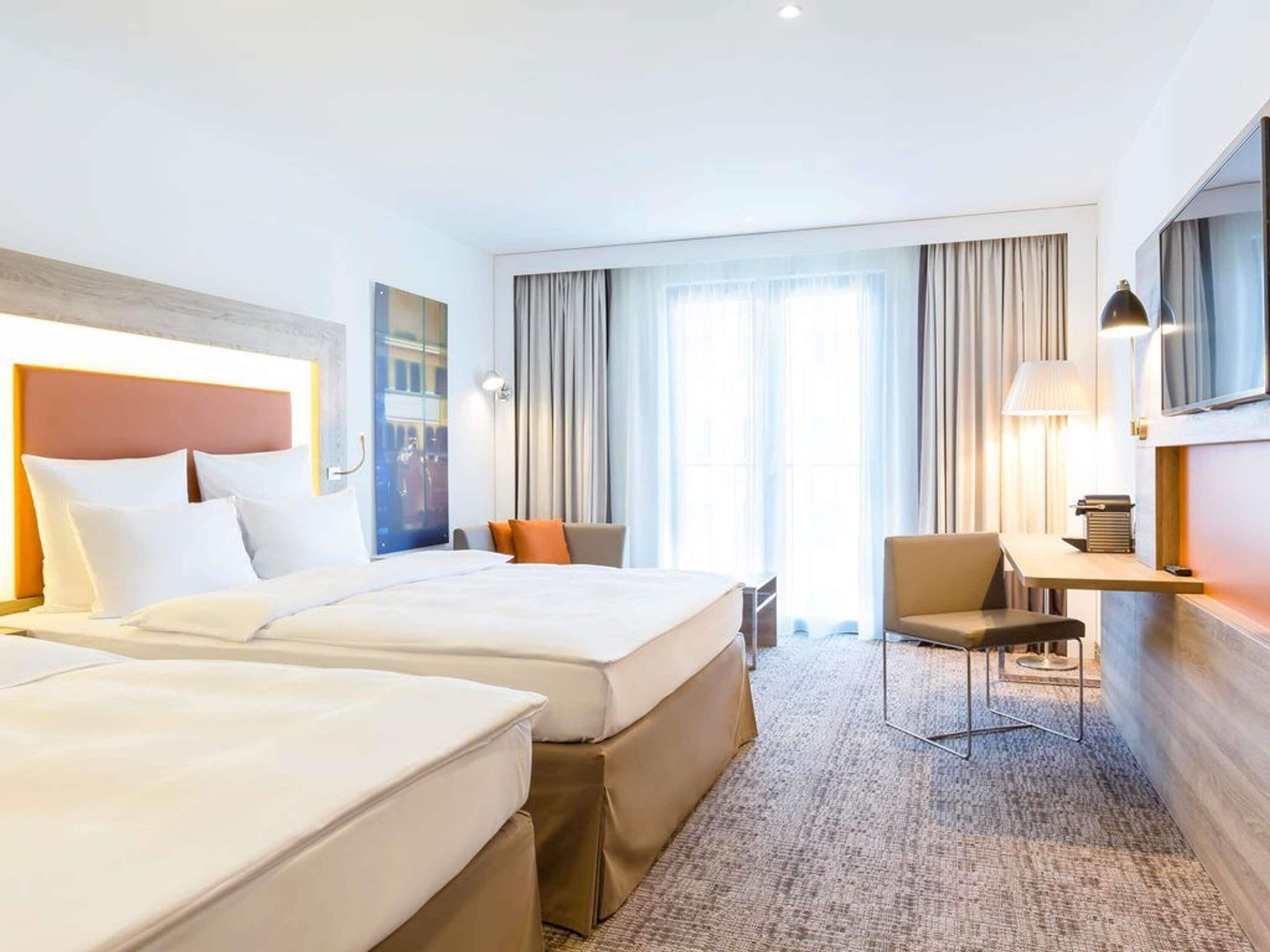A hotel room with two beds, a desk, a chair, and a large window with curtains.