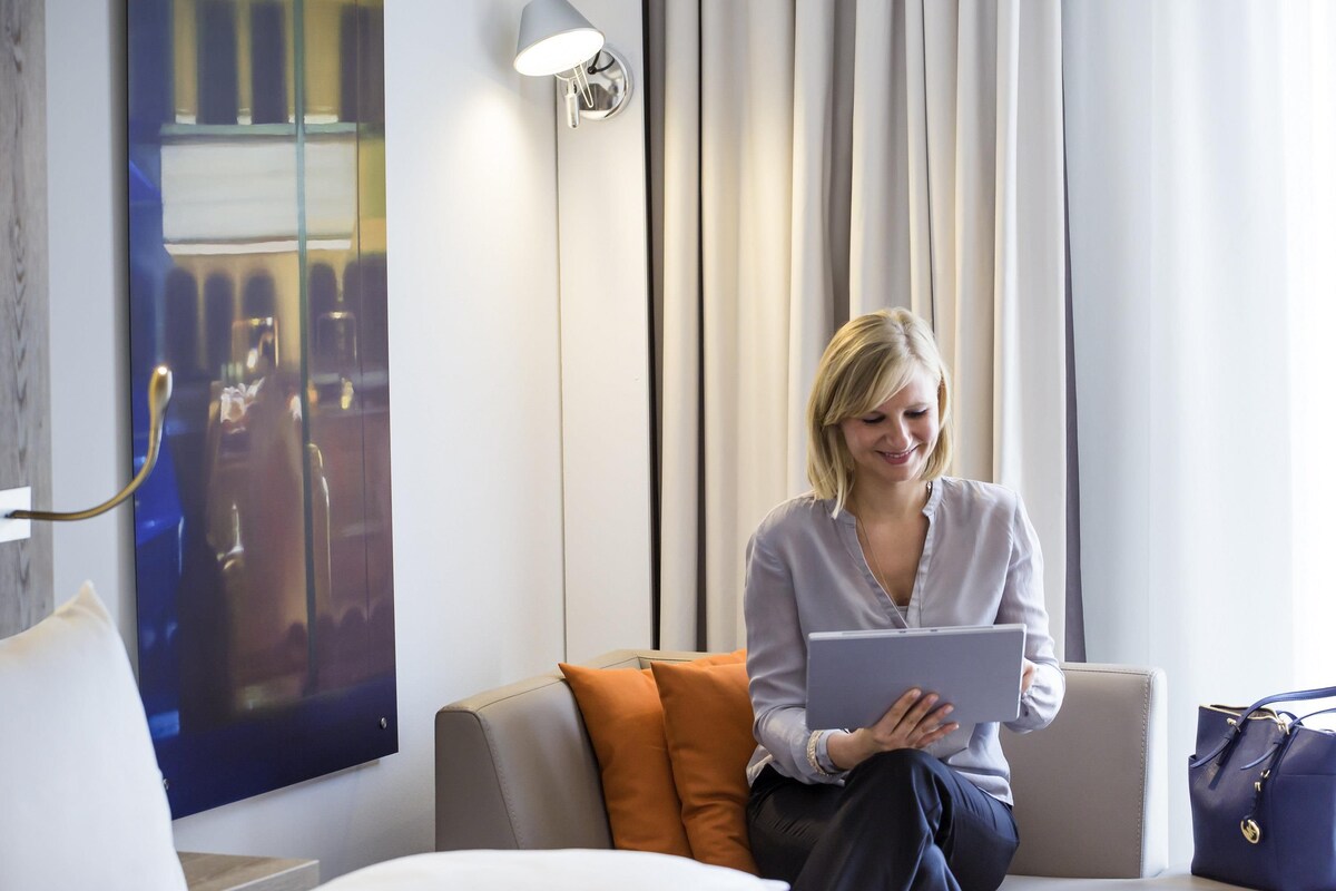 A person sitting on a bed with a laptop, a blue bag, and a pillow.
