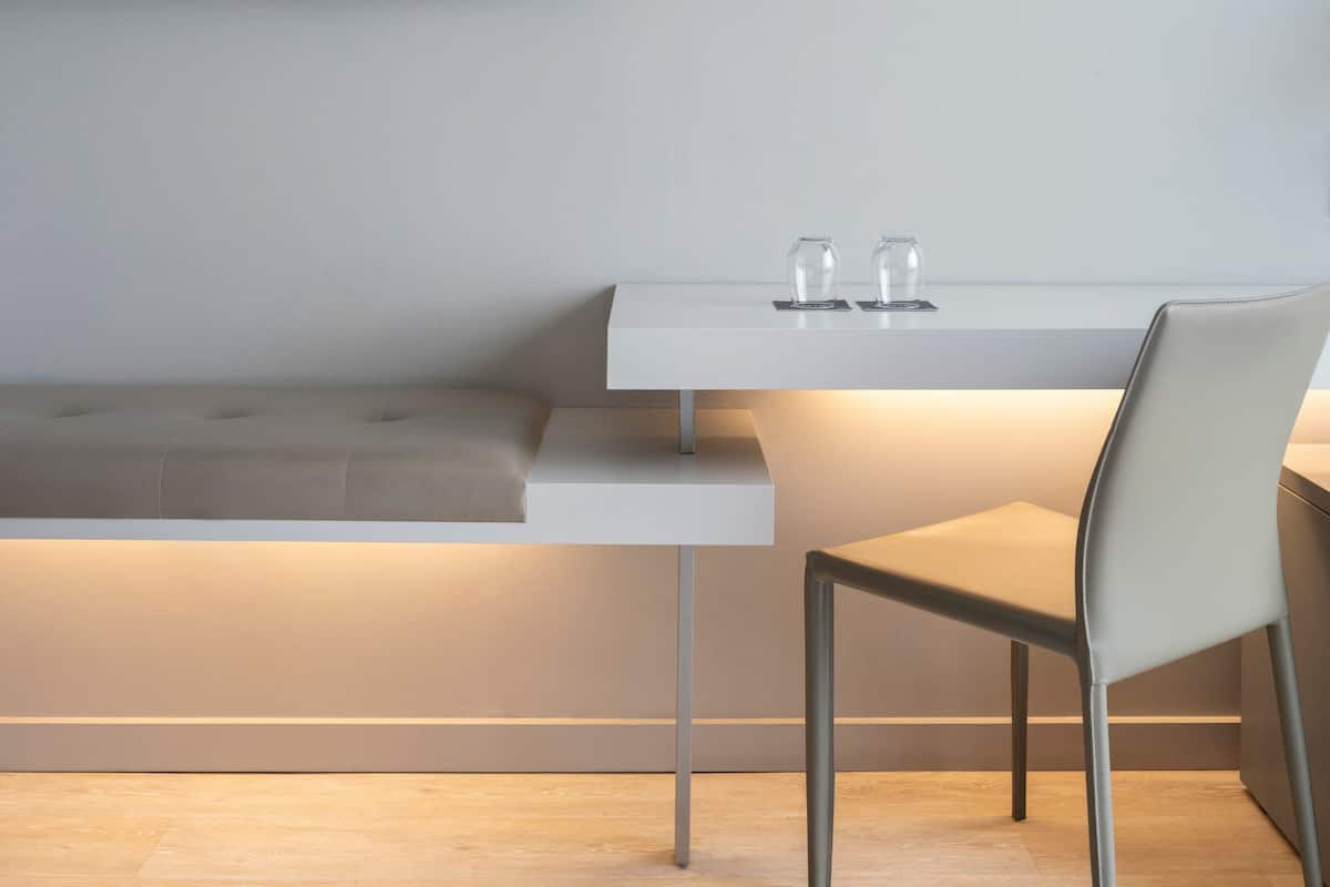A modern, minimalist room with a white bench, a table with two glasses, and a chair.