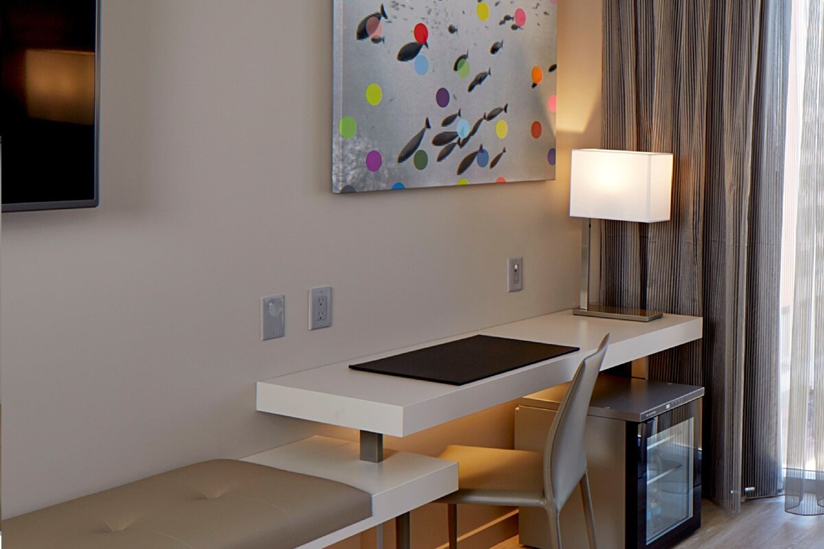 A modern hotel room with a white desk, a gray chair, a bench, a flat-screen TV, a painting, a lamp, and a window with curtains.