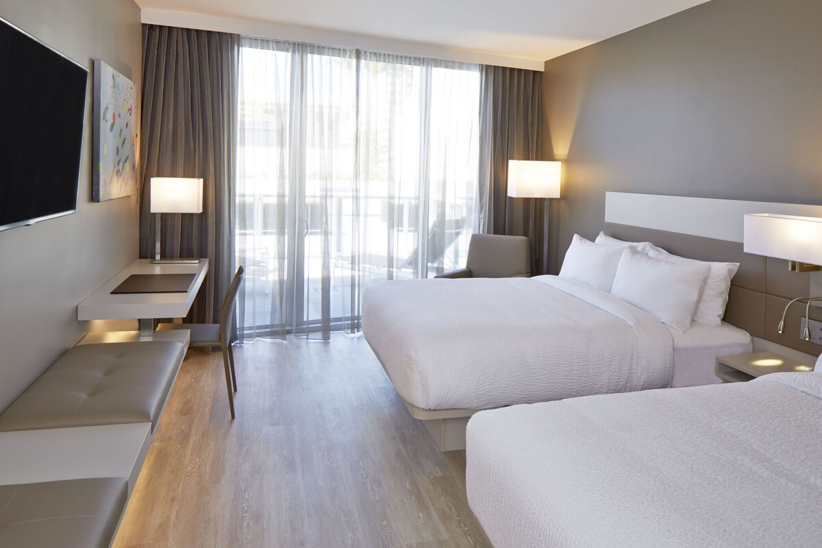 A hotel room with two beds, a desk, and a chair. There is a large window with curtains, a flat-screen TV, and two bedside lamps.